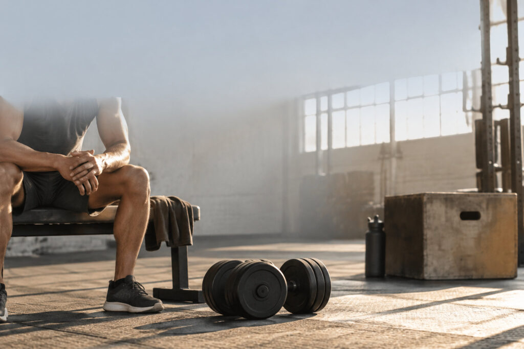 Un athlète assis sur un banc de musculation, se reposant après un entraînement intense, avec des haltères au sol.