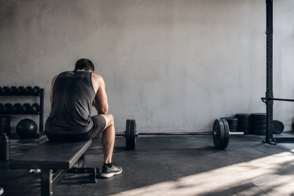 Un homme musclé assis seul sur un banc de musculation, la tête baissée, devant une barre chargée dans une salle de sport.