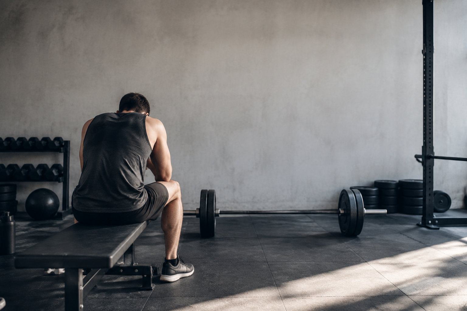 Un homme musclé assis seul sur un banc de musculation, la tête baissée, devant une barre chargée dans une salle de sport.