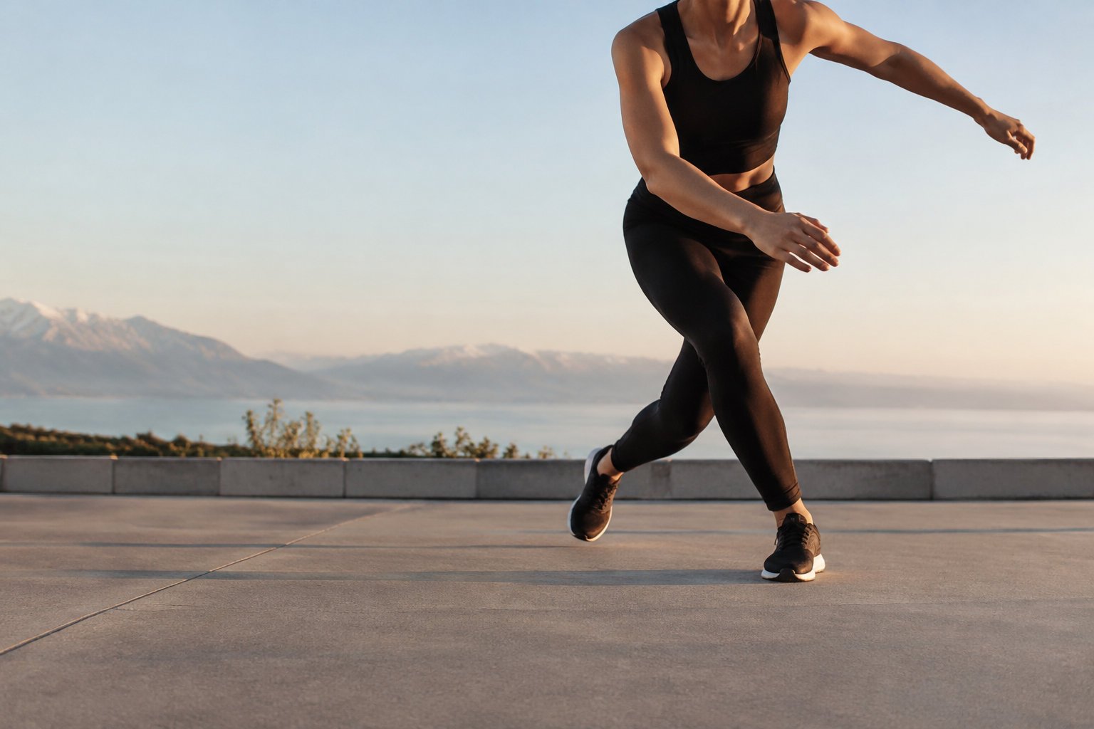 Une sportive en tenue noire effectue un pas du patineur sur une terrasse avec vue sur un lac et des montagnes.